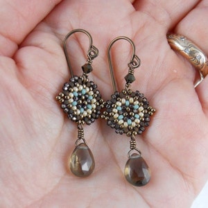 May include: A pair of dangle earrings with a geometric beaded design in shades of brown, beige, and turquoise. The earrings feature a teardrop-shaped smoky quartz gemstone.