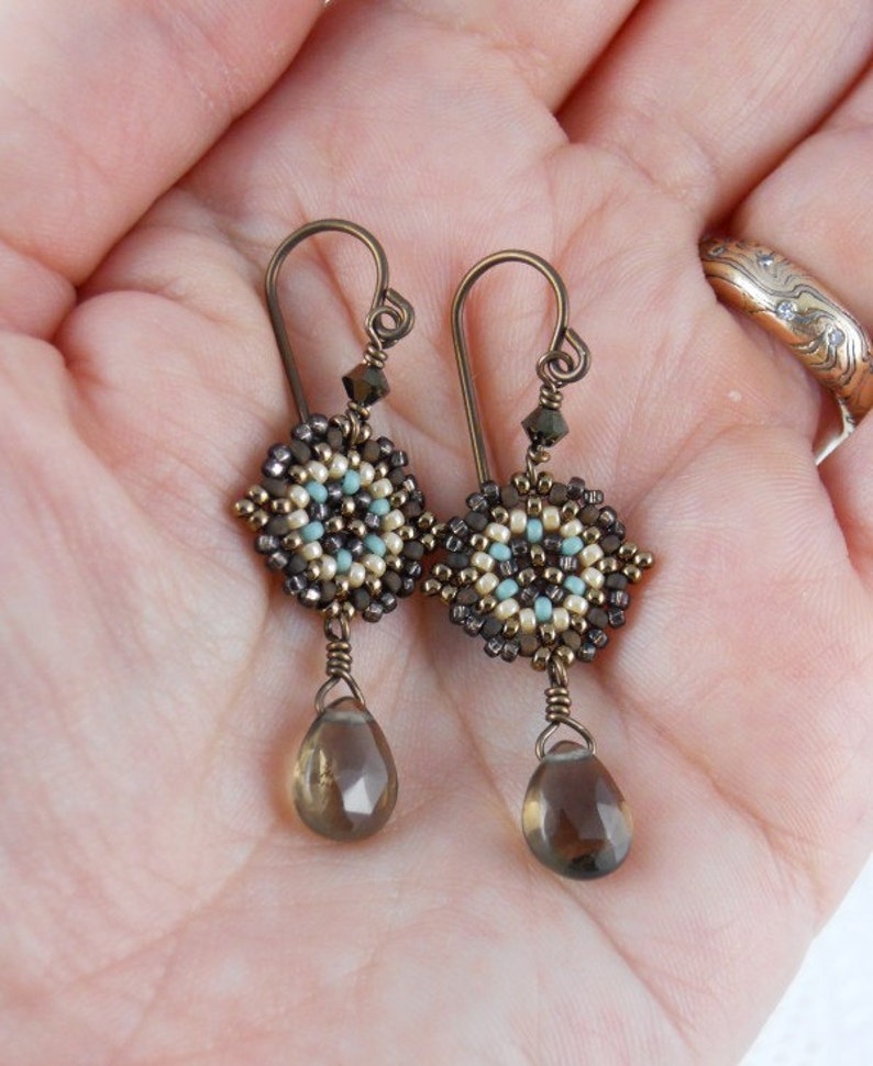 May include: A pair of dangle earrings with a geometric beaded design in shades of brown, beige, and turquoise. The earrings feature a teardrop-shaped smoky quartz gemstone.