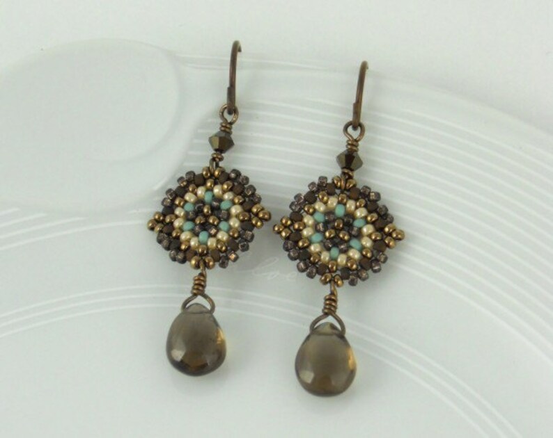 May include: A pair of dangle earrings with a geometric design made of brown, white, and turquoise beads. The earrings have a teardrop-shaped brown gemstone hanging from the bottom.