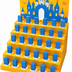 May include: A yellow tiered display stand featuring a blue castle silhouette and pink butterfly cutouts on the back panel. The stand has five levels with multiple circular cutouts, each holding a small, upright, rectangular blue block.