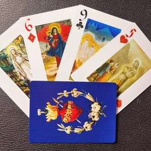 Puede incluir: Un juego de cartas de juego con imágenes religiosas. Las cartas están dispuestas en forma de abanico, con el 6 de diamantes, el 9 de tréboles y el 7 de corazones visibles. Las cartas están decoradas con imágenes de santos y símbolos religiosos.