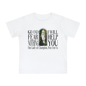 May include: A white short-sleeved t-shirt featuring a central graphic of a stained-glass window depicting a religious figure. The text reads: GO AND FEAR NOTHING I WILL HELP YOU and Our Lady of Champion, Pray For Us in black lettering.
