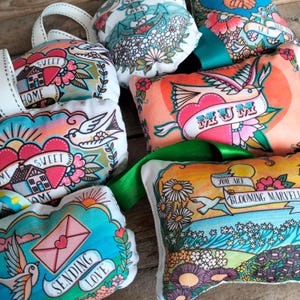 May include: Assortment of colorful fabric pouches with various designs and text, including "Sweet Home", "Mum", "You Are Blooming Marvelous", and "Sending Love".