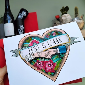 May include: A white card featuring a heart-shaped illustration with a banner reading JESS & IZAAK. The design includes two hands holding each other, surrounded by colorful flowers, leaves, and intricate tattoo-style line work in red and green.
