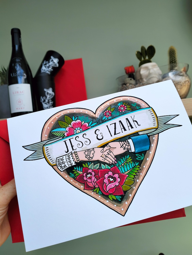 May include: A white card featuring a heart-shaped illustration with a banner reading JESS & IZAAK. The design includes two hands holding each other, surrounded by colorful flowers, leaves, and intricate tattoo-style line work in red and green.