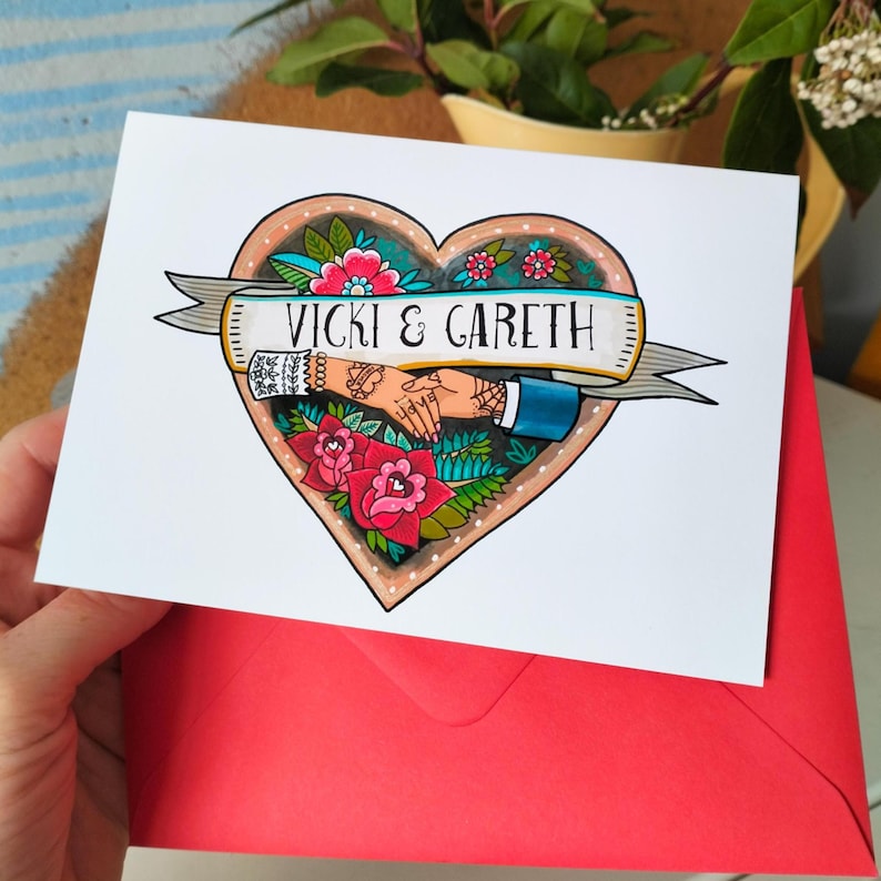 May include: A white greeting card featuring a heart-shaped illustration with a banner reading VICKI & GARETH. The design includes two hands holding each other, surrounded by red and pink flowers with green leaves, all in a tattoo-inspired style.