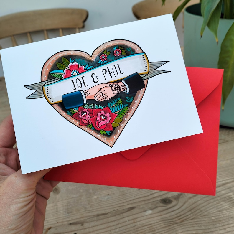 May include: A white greeting card featuring a heart-shaped illustration with a banner across the center that reads JOE & PHIL. The design includes two hands shaking, surrounded by red and pink flowers, green leaves, and a bright red envelope.