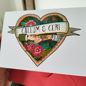 May include: A white greeting card featuring a heart-shaped illustration with red roses, green leaves, and a banner reading CALLUM & CERI. The design depicts two hands holding each other, one with a green sleeve and one with a white lace cuff.