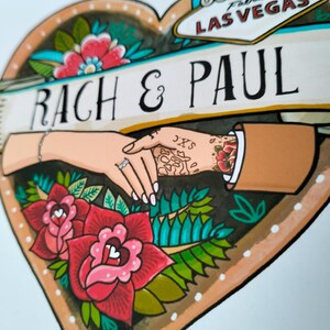 May include: A heart-shaped illustration featuring the text RACH & PAUL and a Las Vegas sign graphic. The design depicts two hands holding each other, one with a ring and bracelet, the other with tattoos, surrounded by red roses and green leaves.