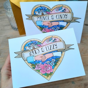 Puede incluir: Dos tarjetas de felicitación con diseños en forma de corazón. Cada tarjeta presenta una pancarta con nombres, un castillo y manos entrelazadas. Las tarjetas están decoradas con flores y una combinación de colores azul y dorado. Una tarjeta dice "Prince & Cindy", la otra "Brad & Lizzy".