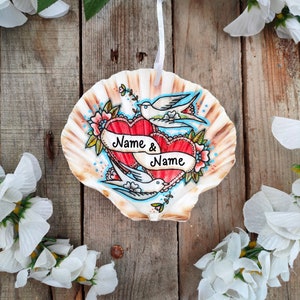 May include: A hand-painted seashell ornament featuring two red hearts with a banner that reads "Name & Name". Two white doves and floral accents surround the hearts. The ornament hangs from a white ribbon.