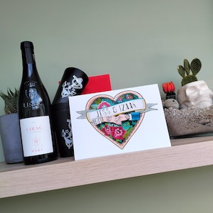 May include: A rectangular greeting card featuring a colorful heart-shaped illustration with two hands holding each other, red roses, and a banner with the text JESS & IZAAK. The card is positioned next to a black wine bottle and a black tumbler.