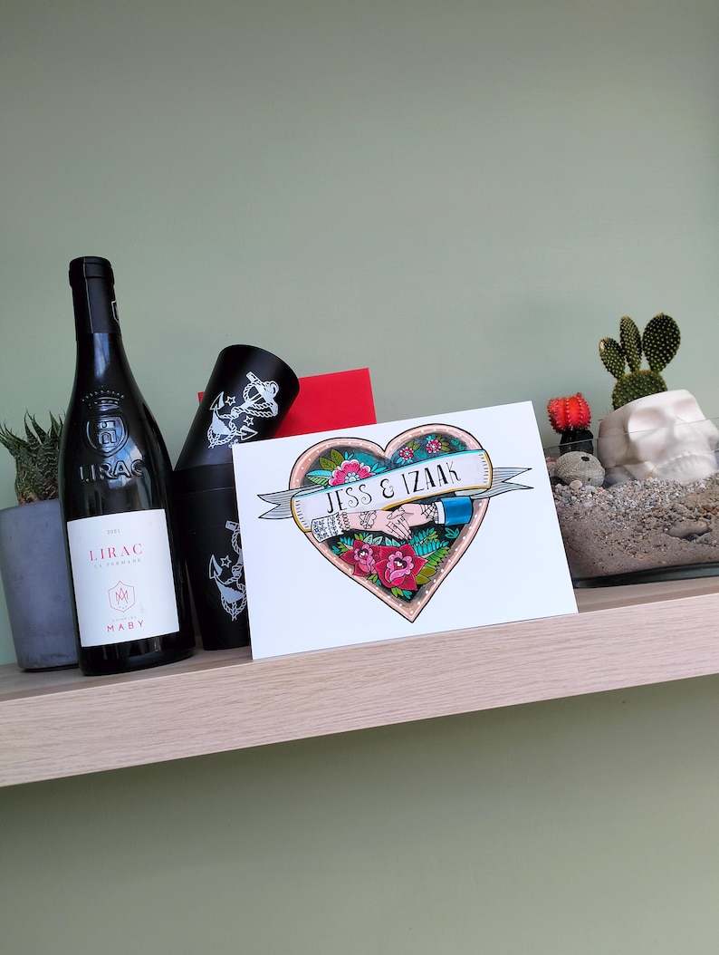 May include: A rectangular greeting card featuring a colorful heart-shaped illustration with two hands holding each other, red roses, and a banner with the text JESS & IZAAK. The card is positioned next to a black wine bottle and a black tumbler.