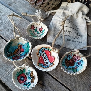 May include: A collection of hand-painted seashell decorations with twine loops. The shells are decorated with various designs, including a bird, flowers, and a heart with the word "LOVE". A small bag with "Stormy Seas UK Brighton" is also visible.