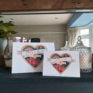 May include: Two white rectangular cards featuring a heart-shaped illustration with red flowers, green leaves, and a banner. One card displays the text EMILY & JASON, while the other displays STEPH & LEE, both showing two hands holding each other inside the heart.