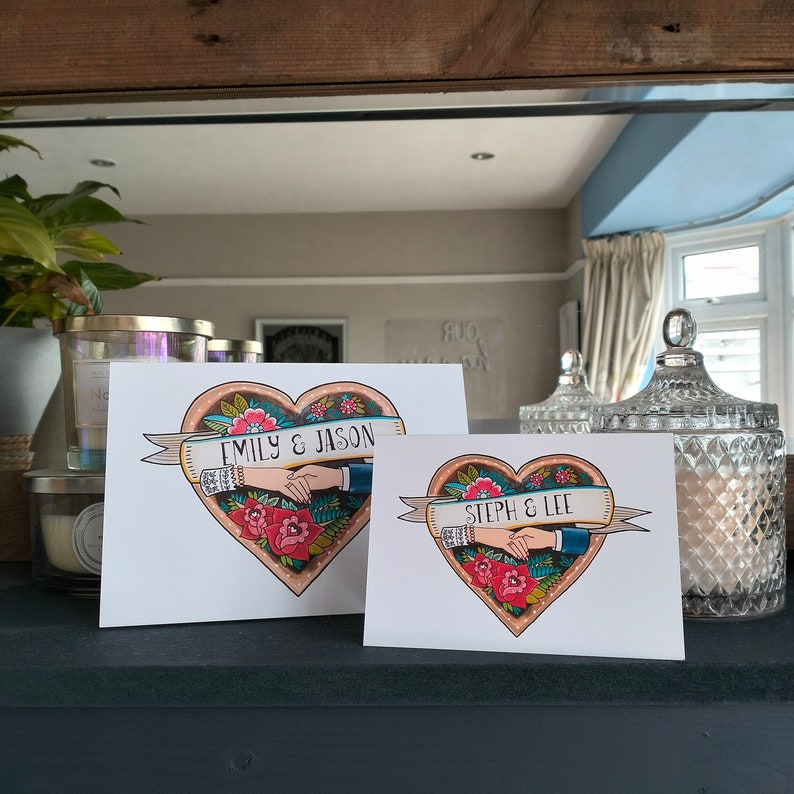 May include: Two white rectangular cards featuring a heart-shaped illustration with red flowers, green leaves, and a banner. One card displays the text EMILY & JASON, while the other displays STEPH & LEE, both showing two hands holding each other inside the heart.