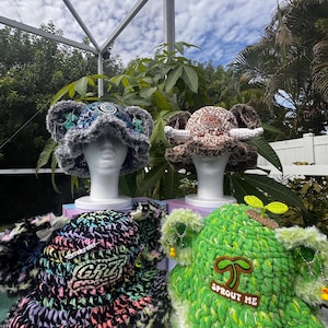 May include: Four plush bucket hats with animal ears. Top left is blue and grey with a spiral patch. Top right is brown with white tusks. Bottom left is black with neon accents and a GRiZ patch. Bottom right is green with a sprout and SPROUT ME patch.