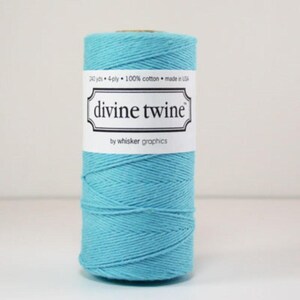 May include: A spool of turquoise Divine Twine by Whisker Graphics. The label states it is 100% cotton, 4-ply, and contains 219 metres. The twine is a vibrant blue-green colour, tightly wound around a cardboard core.