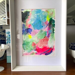 May include: A framed abstract painting with bright colors, including pink, blue, green, and yellow. The painting is in a white frame and is on a wooden surface.