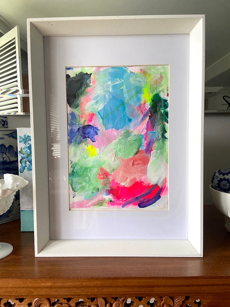 May include: A framed abstract painting with bright colors, including pink, blue, green, and yellow. The painting is in a white frame and is on a wooden surface.