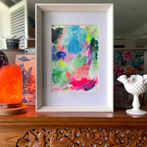 May include: A framed abstract painting with bright colors, including pink, blue, green, and yellow. The painting is in a white frame and is sitting on a wooden table.