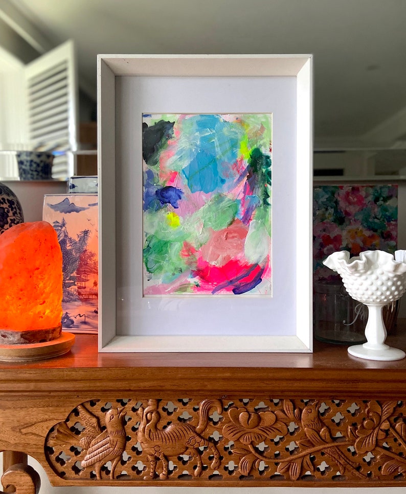 May include: A framed abstract painting with bright colors, including pink, blue, green, and yellow. The painting is in a white frame and is sitting on a wooden table.