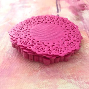 May include: A stack of bright pink, circular paper doilies. Each doily features a delicate, floral lace pattern around the edge and a textured center. The doilies are stacked on top of each other, creating a layered effect.