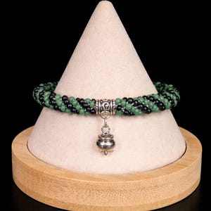 May include: A beaded bracelet with alternating green and black beads. The bracelet has a silver-colored clasp and a small, decorative charm. The bracelet is displayed on a white cone-shaped stand with a wooden base.