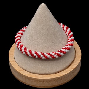 May include: A red and white beaded bracelet on a wooden display stand. The bracelet is made of small, round beads that are arranged in a spiral pattern.