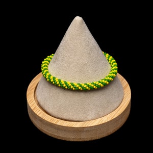 May include: A green and yellow beaded bracelet on a gray and brown display stand.