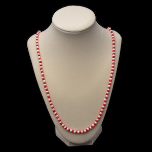 May include: A red and white beaded necklace on a white display stand. The necklace is made of small, round beads.