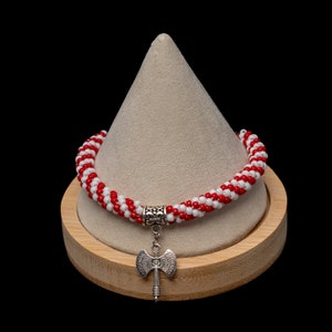 May include: Red and white beaded bracelet with a silver axe charm. The bracelet is displayed on a wooden stand.