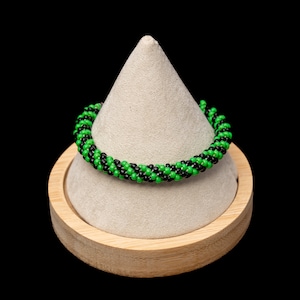 May include: A beaded bracelet with alternating rows of black and green beads. The bracelet is displayed on a light beige cone-shaped stand, which is set on a round wooden base. The background is solid black.