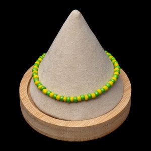 May include: A beaded bracelet with alternating yellow and green beads. The bracelet is displayed on a light tan, cone-shaped stand, set on a round, wooden base. The background is black, highlighting the bracelet's colors.