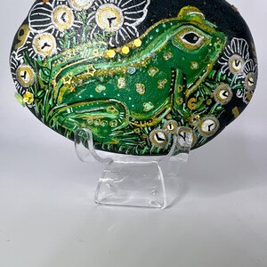 May include: Hand-painted rock art featuring a green frog with gold accents, surrounded by white flowers and gold clock faces on a black background. The rock is displayed on a clear plastic stand.