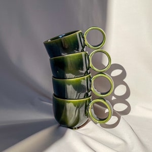 Peut inclure: Une pile de quatre tasses en céramique verte avec des poignées arrondies. Les tasses sont empilées en forme de pyramide, la plus grande tasse en bas et la plus petite tasse en haut.
