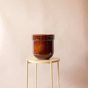 Può includere: Un grande vaso di ceramica marrone lucido con un bordo largo, appoggiato su un piccolo tavolino rotondo in metallo giallo. Il vaso ha un colore marrone scuro intenso e una superficie liscia e riflettente. Il tavolo ha un design semplice e minimalista.