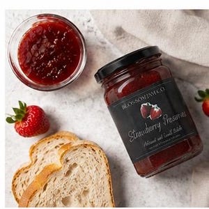 May include: A jar of strawberry preserves with a black label and the text "Strawberry Preserves". A small glass bowl of preserves, two slices of bread, and a fresh strawberry are also visible.