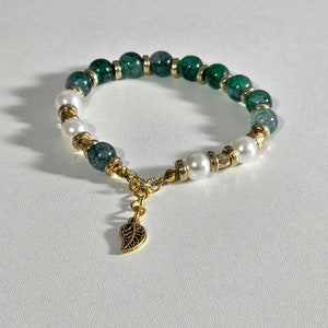 Könnte beinhalten: Ein grünes und weißes Perlenarmband mit einem goldenen Blattanhänger. Das Armband besteht aus grünen und weißen Perlen und hat eine goldene Kette mit einem Blattanhänger.