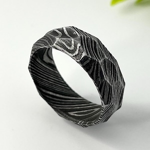 May include: A faceted ring crafted from dark Damascus steel, featuring a distinct layered pattern of black and silver tones. The band has a multi-faceted, angular exterior surface that creates a textured appearance throughout the metal.
