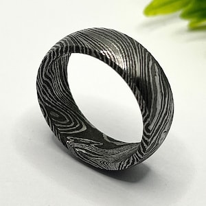 Puede incluir: Un anillo de acero de Damasco con un patrón arremolinado único. El anillo tiene un aspecto oscuro y metálico y un acabado liso y pulido. La banda mide aproximadamente 0,8 cm de ancho. El anillo está sobre un fondo blanco con follaje verde.