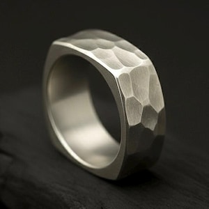 May include: A silver ring with a hammered texture and a square shape. The ring is set against a dark background, highlighting its metallic sheen and unique design. The band has a textured surface.