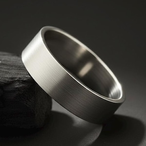 May include: A brushed silver ring with a flat band. The ring is set against a dark background, highlighting its metallic sheen. The ring's simple design and polished finish suggest a modern aesthetic.