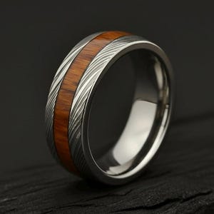 May include: A silver ring with a wood inlay is shown in close-up. The ring features a polished silver band with a central inlay of reddish-brown wood. The wood grain is visible, and the ring is set against a dark background.