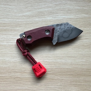 May include: A small, dark gray pocket knife with a burgundy wood handle and a red lanyard. The knife has a textured blade and a lanyard loop.