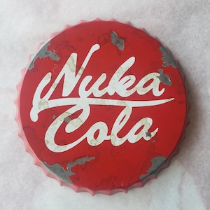 May include: A red metal bottle cap featuring the white text "Nuka Cola". The cap has a distressed, aged look with areas of paint loss, suggesting a vintage style. The edges are crimped and the overall design is circular.