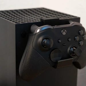 Può includere: Una console Xbox Series X nera con un controller Xbox nero collegato. Il controller è appoggiato in un supporto nero attaccato alla console. La console è un cubo nero con una parte superiore strutturata.