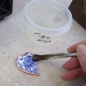 May include: A close-up of a blue and white ceramic heart-shaped piece being brushed with flux. The flux is in a clear plastic container labelled "Gel Flux".
