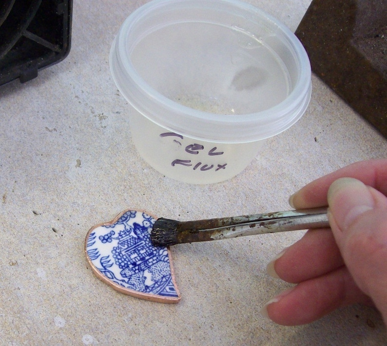 May include: A close-up of a blue and white ceramic heart-shaped piece being brushed with flux. The flux is in a clear plastic container labelled "Gel Flux".