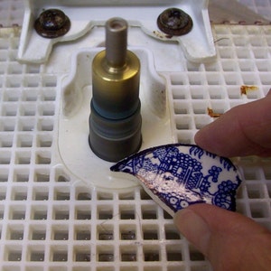 May include: A close-up of a broken piece of blue and white china being held by a hand. The china is being held over a white gridded surface with a metal tool in the centre.